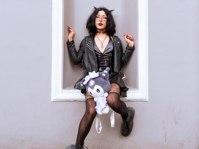 NoemiSky with black cat ears, leather jacket, corset, fishnet stockings, black boots, holding a plush grey cat toy, sitting in a white window recess, red lipstick, dark makeup.
