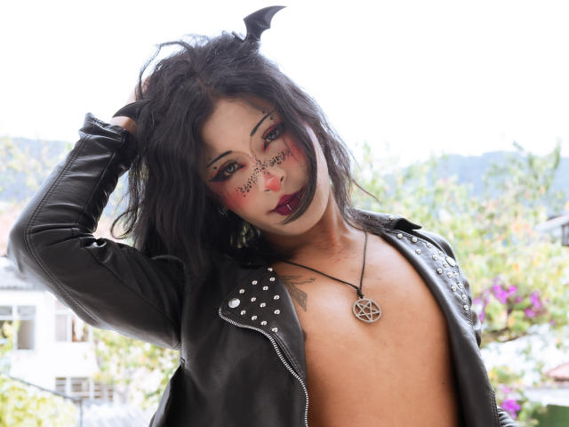 NoemiSky is a woman with dark hair and bat ears, wearing a black leather jacket with silver studs, and no shirt. She has black lipstick, red face paint with black lines, and a pentagram necklace. Her right hand is in her hair. The image is a photograph with natural light. She has a tattoo on her left side. Her skin is light brown. Her expression is neutral. The background is blurred with greenery and a house. The image has a watermark in the bottom right corner.