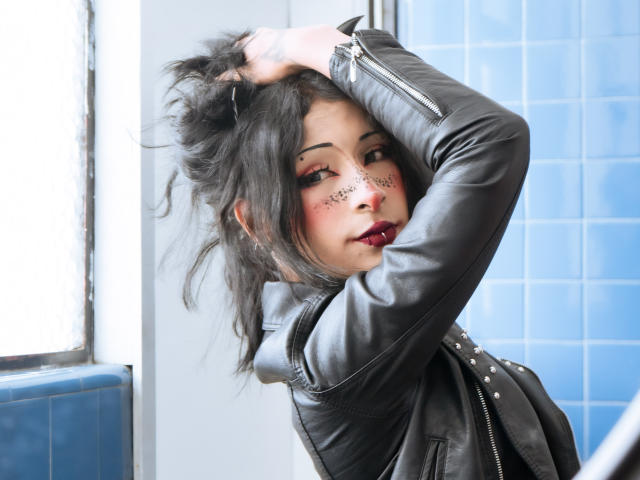 NoemiSky with dark hair, red makeup, black leather jacket, raising her hand to her messy hair, looking sideways with red lipstick, blue tiled background.