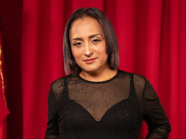 AmandaSolery with medium brown skin, straight dark brown hair, wearing a sheer black dress with sparkly dots, showing her small tits, and a slight smile. Red curtain background.