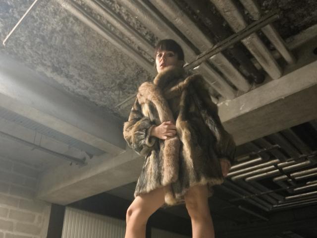 OrianaLaFrancaise stands in a dark, gritty basement wearing a fluffy, brown and black fur coat, legs exposed, holding the coat with one hand, looking up.