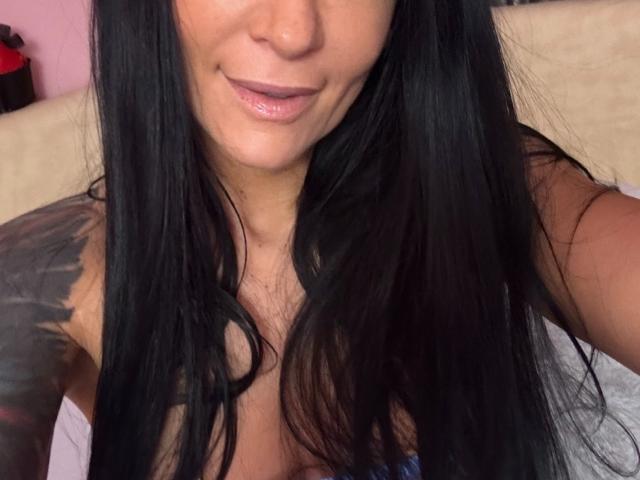 AndraD has long black hair, a tattoo on her right shoulder, and is smiling with shiny pink lips. Her small, shiny tits are visible. AndraD is taking a selfie.
