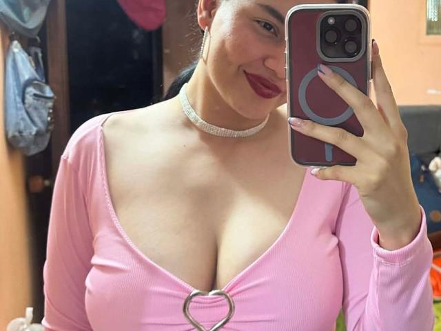 JulietaFoster69 with big tits, pink ribbed top, heart ring, choker, smiling, holding phone, pink nails, red lipstick, brown hair in ponytail, mirror selfie, iPhone.
