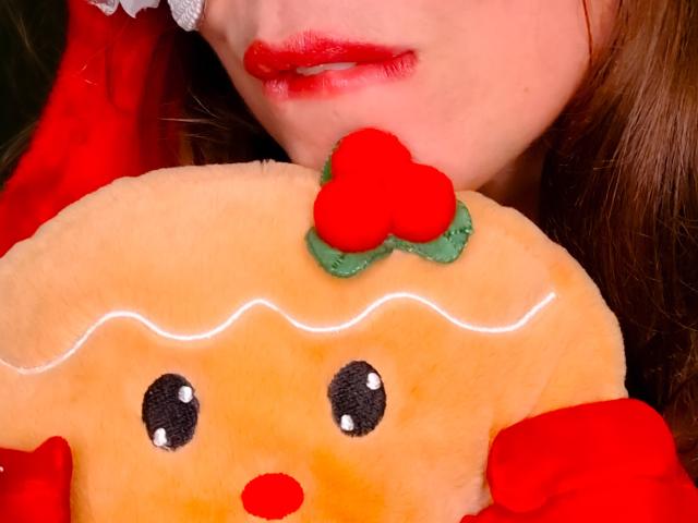 JuliaFrancaise's face with red lipstick, holding a cute, orange, plush toy with black eyes, red nose, and green bow with a red heart, against a dark background.