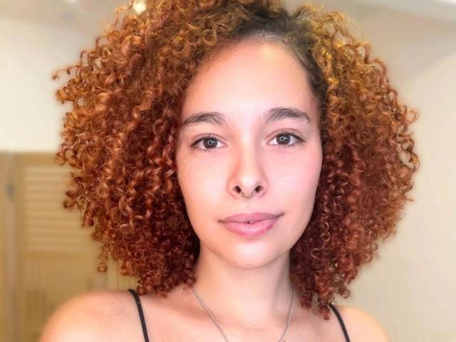 LizzaMonroe-hot has light brown curly hair, a cute face, and is wearing a black spaghetti strap top. She has a subtle smile and a silver necklace.