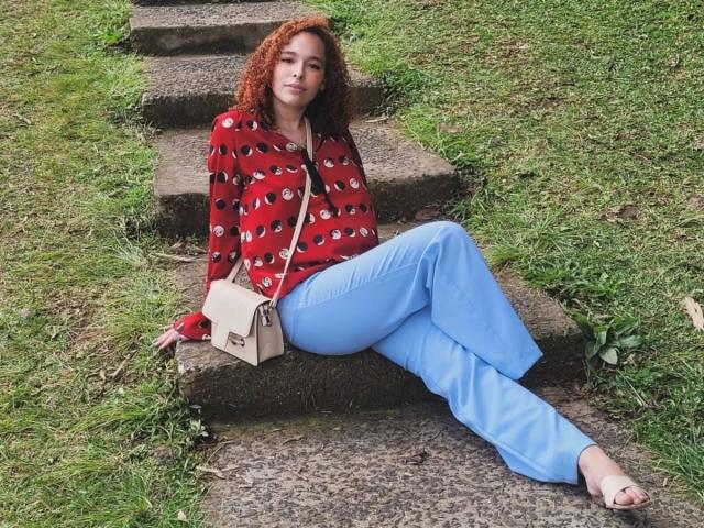 LizzaMonroe-hot with curly red hair, red eye-pattern shirt, blue pants, beige bag, and open-toe sandals, sitting on stone steps, looking relaxed.