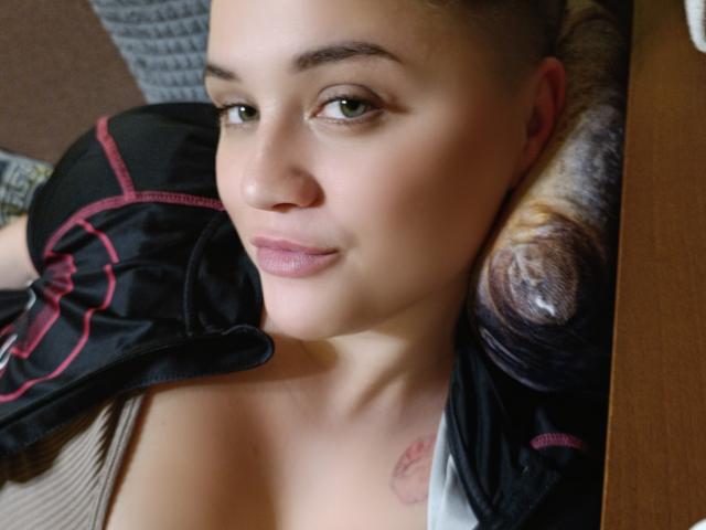 TessaxRenata with light skin and green eyes, lying down, black and red jacket over shoulder, small tattoo on chest, brown hair in bun, slight smile, soft pink lipstick.