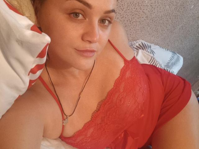 TessaxRenata in red lace camisole, lying on bed, showing small tits, slight cleavage, necklace with star pendant, looking at camera with a subtle smile.