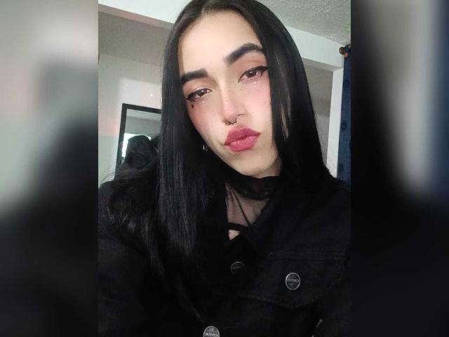 SamiSmith69 has long black hair, dark eyebrows, pink eyeshadow, nose ring, and red lips. She's wearing a black button-up shirt with a sheer neckline.