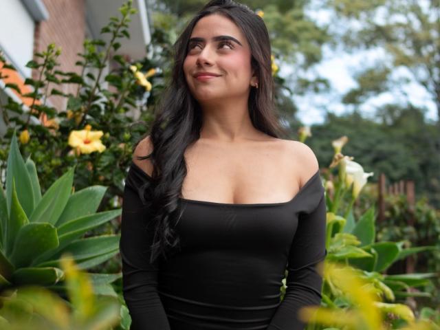 MaddieColins with long black hair, wearing an off-shoulder black dress, showing her boobs, standing in a garden with yellow flowers and green leaves, smiling slightly.