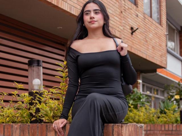 MaddieColins with dark hair, black off-shoulder dress, and black pants, sitting on brick ledge, holding strap, showing her tits slightly, confident expression, outdoor setting, green plants.
