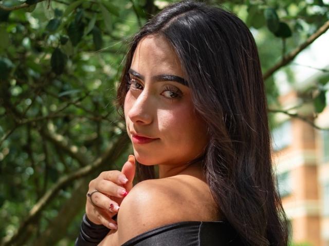 MaddieColins with long dark hair, looking over shoulder, black off-shoulder top, red nails, slight smile, brown eyes, soft light, green tree background, right hand touching chin.