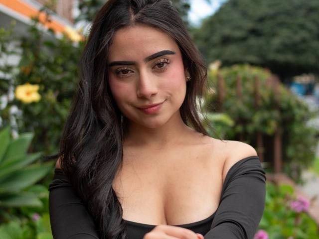 MaddieColins has long dark hair, a cute face, and is wearing an off-shoulder black top that shows her big boobs. She's smiling slightly and looking at the camera.