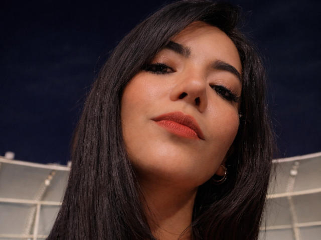 SofieWhite's face with long dark hair, slightly upturned nose, full lips with orange lipstick, and subtle makeup. She's wearing small hoop earrings and looking down with a slight smile.