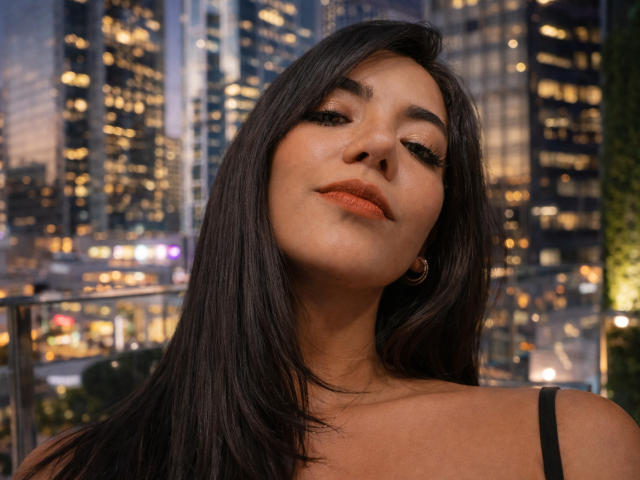 Close-up of SofieWhite with straight, dark brown hair, slightly parted to reveal her face. She has a light olive skin tone and full lips with orange lipstick. Her eyebrows are thick and slightly arched. She's wearing a black spaghetti strap top. Her head is tilted slightly back, eyes half-open, looking upwards. She's wearing small hoop earrings. The background is a cityscape at night with tall buildings and bright lights, creating a blurred, bokeh effect. The image has a soft, warm light that highlights her facial features. Her expression is calm and slightly confident.