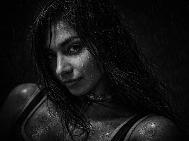 Black and white photo of SofieWhite with wet, dark, tangled hair. Her face is glistening with raindrops. She has dark, intense eyes and a subtle, almost defiant smile. Her wet, dark tank top clings to her skin, highlighting her shoulders and collarbone. The texture of her hair and skin is detailed, showing individual strands and droplets. The overall composition is dark and moody, with a focus on SofieWhite's expressive face and wet appearance. The photograph emphasizes contrast and texture.