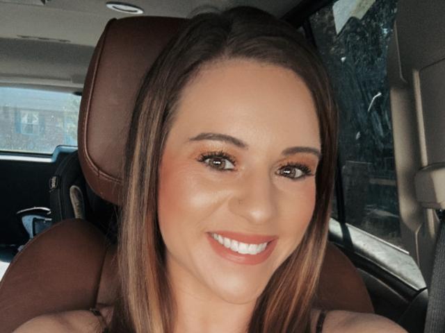 Rubie69's smiling face with brown eyes, long brown hair, and red lipstick, sitting in a car, showing off her cute nose and full tits. She's looking at the camera.