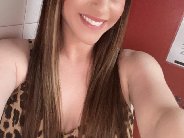 Rubie69 with long brown hair, smiling, showing white teeth, wearing a leopard print top, and a necklace, in a selfie. Her tits are partially visible. Red wall in background.