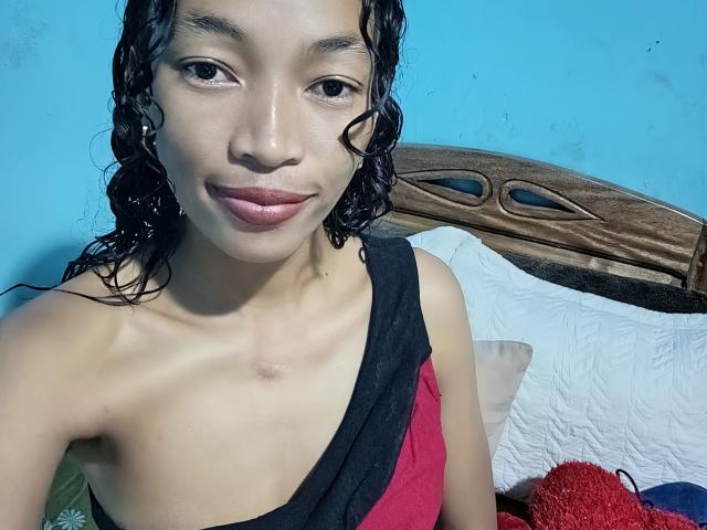 RedBellah has light brown skin, curly black hair, and a slight smile. She's wearing a red and black one-shoulder top, showing her bare shoulders and chest.