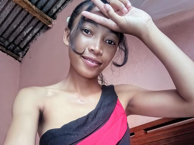 RedBellah with small tits, dark hair, and brown skin, wearing a black and pink one-shoulder top, smiling, making a peace sign with her right hand.