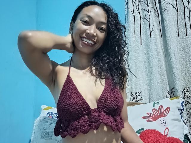 RedBellah with curly black hair, smiling, wearing a maroon crochet bikini top, showing her small tits, hand behind head, blue wall, white curtain with black tree pattern.