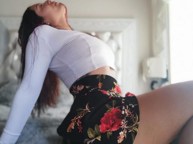 LaurenSexyX leans back, eyes closed, head tilted up. She's wearing a white long-sleeve crop top and a black floral skirt with red roses. Her dark hair is loose, and she's arching her back, showcasing her midriff. The white headboard in the background is blurred. Light from the right side casts soft shadows. Her skirt is tight, highlighting her thighs. The composition is slightly diagonal, emphasizing her posture. The texture of her top is smooth, contrasting with the floral pattern of her skirt. The image is focused on LaurenSexyX's upper body and legs.