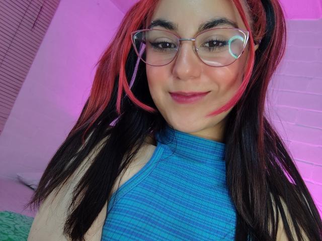 AmyStreetwise69 with red and black hair, glasses, blue ribbed top, smiling, pink and purple lighting, visible nose, and subtle makeup, showing her face and upper chest.