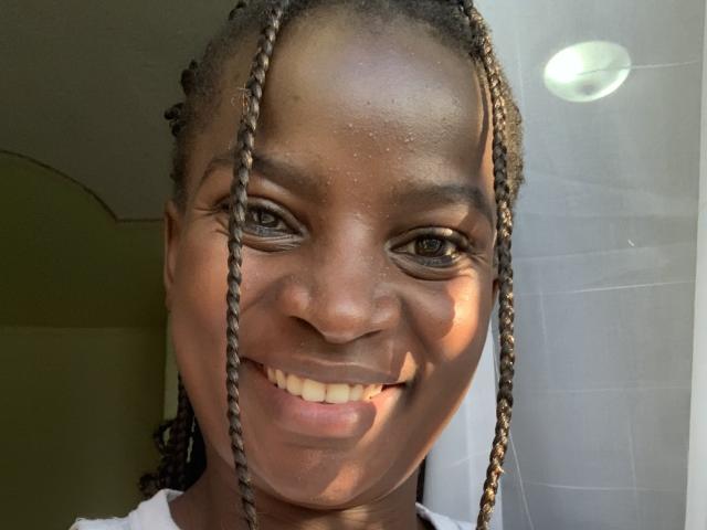 JennyNorah with dark brown skin, long braided hair, smiling, showing teeth, small nose, brown eyes, white shirt, sunlight highlighting face, gentle shadow on right side.