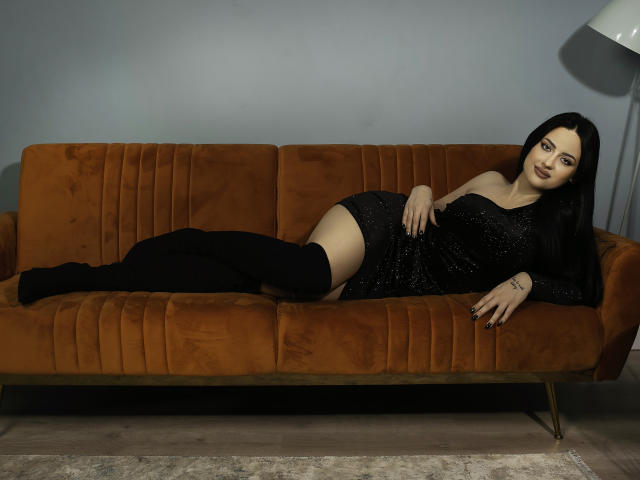 VanessaEsther is lying on an orange suede couch, wearing a black sparkly dress and black thigh-high socks, with her ass slightly raised and her tits visible.