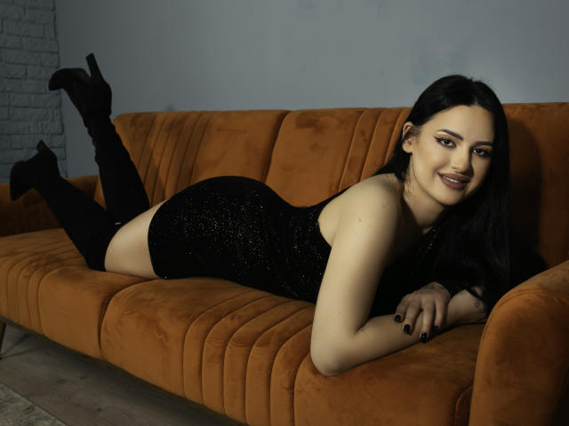 VanessaEsther is lying on an orange suede sofa, wearing a sparkly black dress and black high heels, with her ass raised and a smile on her face.