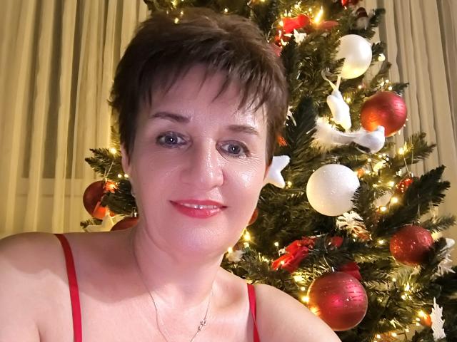 DoraBlueEyess con cabello castaño corto, piel brillante, lápiz labial rojo y tirantes rojos de espagueti, sonriendo frente a un árbol de Navidad con adornos rojos y blancos y luces brillantes..
