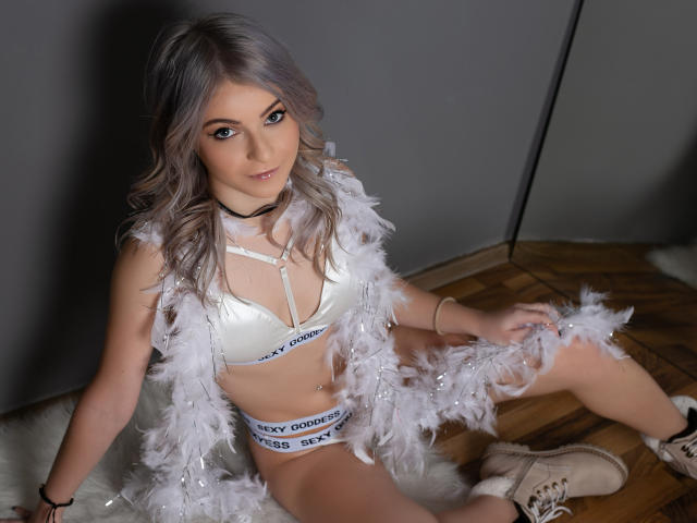 SelenaBellax with silver hair, wearing a white bra and panties, a feather vest, and boots, sitting on a fluffy rug, looking up with a slight smile. Her tits are small.