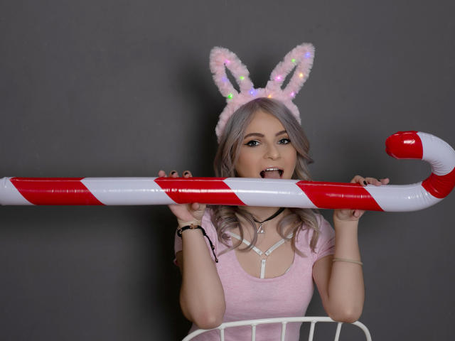 SelenaBellax with pink hair, white furry bunny ears, holding a giant candy cane, tongue out, wearing a pink shirt, gray background, and black bracelet, looking cute and playful.