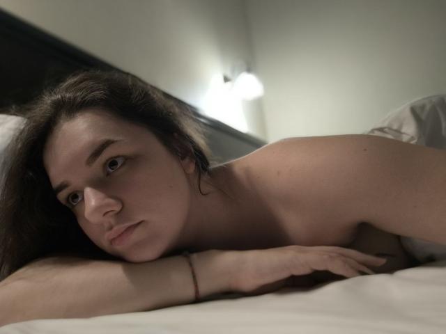 Dreia is lying topless in bed, her bare tits and ass partially covered by white pillows. She has brown hair, a bracelet on her wrist, and a serious expression.
