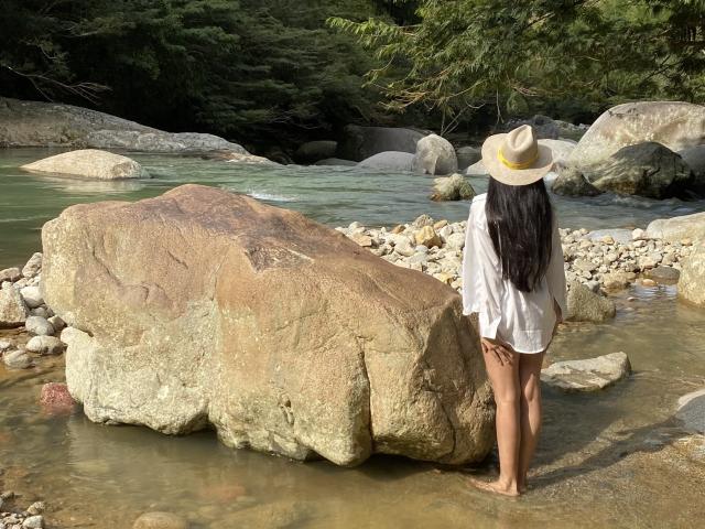 TiffannyRose está descalza junto a una roca, vestida con una camisa blanca, sin pantalones y un sombrero beige, con el pelo largo y negro, frente a un río rocoso. Se puede ver su trasero..