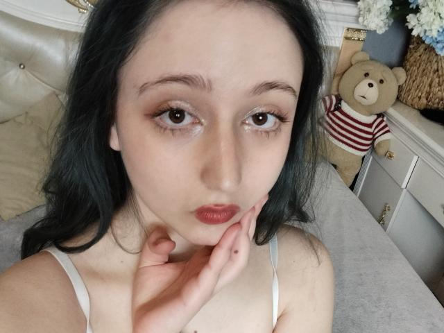 TwinkyKiss with pale skin, dark green eyes, and red lipstick, touching her chin with a hand, wearing a white spaghetti strap top, black hair, and a cute teddy bear.
