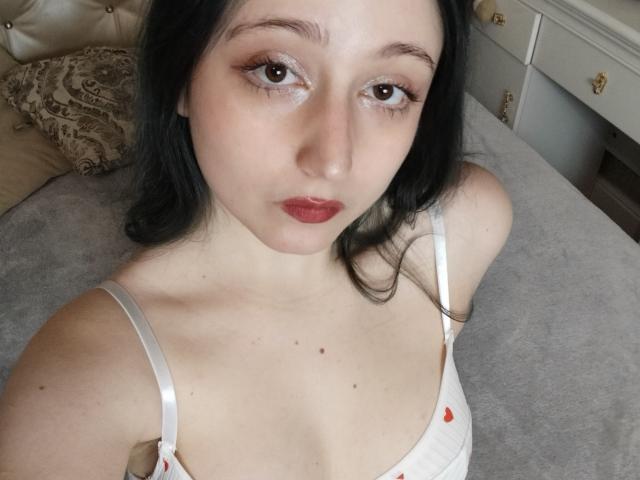 TwinkyKiss has pale skin, dark hair, and red lipstick. She's wearing a white spaghetti strap top with small red patterns, showing her small tits and a few moles on her chest.