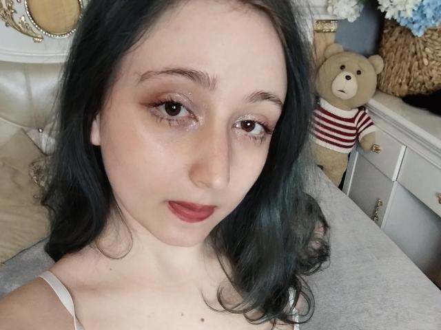 TwinkyKiss has pale skin, dark green wavy hair, and red lipstick. She's wearing a white top, with a cute teddy bear in a red striped shirt behind her.