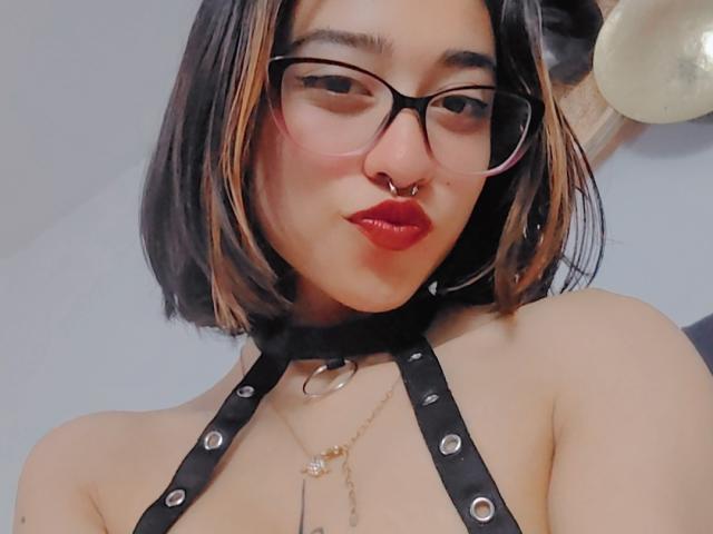 CamiVargas with black bob, pink-tinted glasses, red lipstick, nose ring, black leather harness, necklace, and glasses. Boobs visible, no ass. Light skin, cute, sexy.