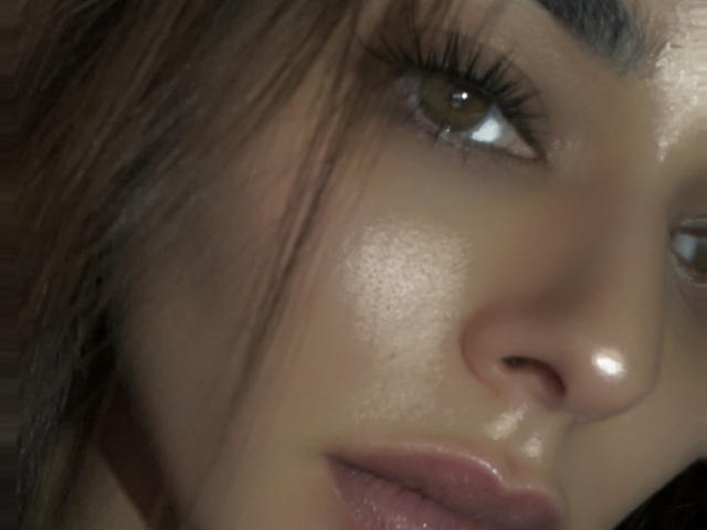 Bibinaa69's shiny face, brown eyes, and full lips with slight gloss. Her long eyelashes and subtle makeup highlight her smooth, slightly tanned skin. Her nose is centered.