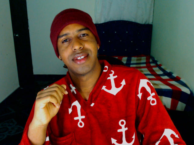 MrJhon69 wearing a red beanie and red shirt with white anchor patterns, smiling with brown eyes, short black hair, and a gold earring, holding his right hand up.