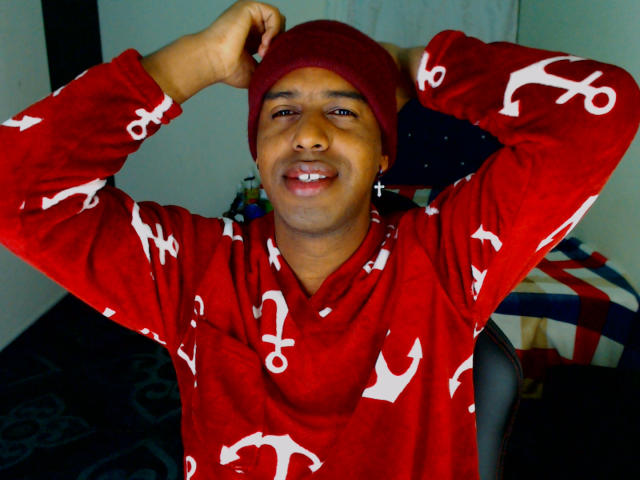 MrJhon6 is in a red hoodie with white anchor patterns, wearing a matching red beanie. He's smiling with his arms raised, hands behind his head. His dark brown skin contrasts with the bright red of his clothing. The background includes a blurred American flag and a dark carpet with a pattern. The image is sharply focused on MrJhon6, with a casual, relaxed atmosphere. The red hoodie is textured, adding depth to the fabric. His confident, cheerful expression is central to the image.