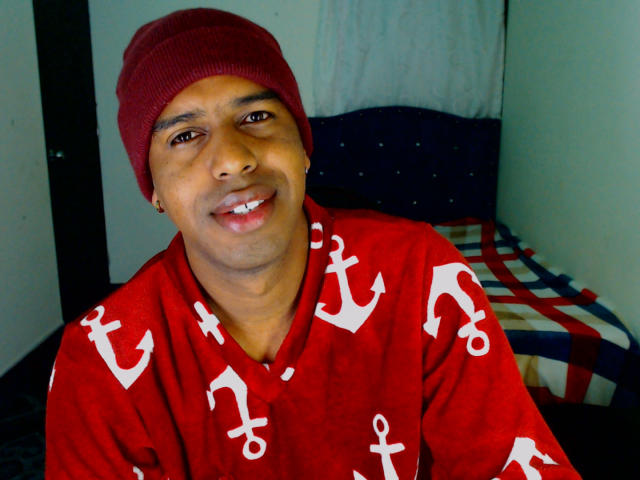 MrJhon69 with brown skin, smiling, wearing a red beanie and red shirt with white anchor patterns, sitting on a checkered bed. His ass is not visible.