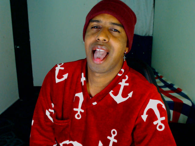 MrJhon6 is a medium-skinned man with a round face and dark eyebrows, wearing a red beanie and a red shirt with white anchor patterns. He's sticking his tongue out with an open-mouthed, playful expression. He has small, round earrings in both ears. The image is brightly lit with a slight shadow on his left side. The background is a simple room with a dark door on the left and a bed with a blue and red patterned blanket on the right.