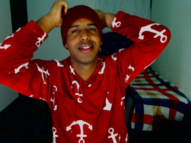 MrJhon69 is a chubby dude with dark skin, smiling with hands on red beanie, wearing red pajamas with white anchors, in a room with a checkered blanket.