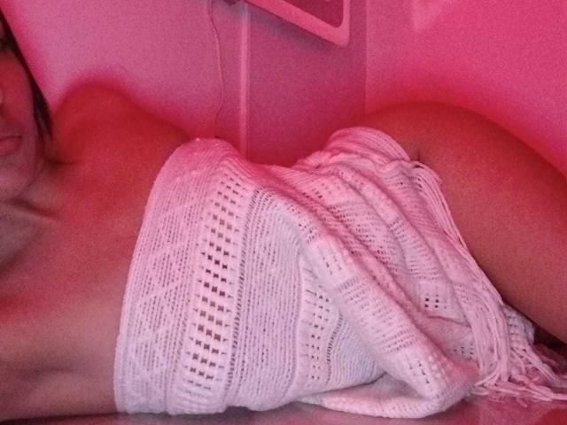 NicolleGlory's bare ass is covered by a white, knitted, fringed dress, lying on a pink-lit surface, showing her tan skin and side profile.