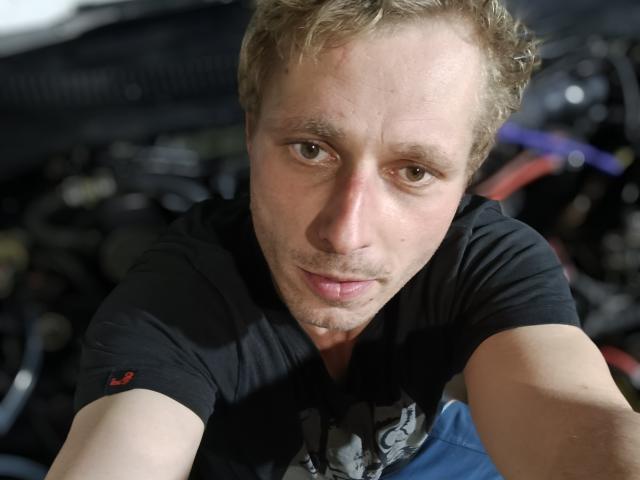 TomTechh with messy blonde hair, light brown eyes, slight beard, black shirt, and subtle grin, leaning forward, looking at camera. Background is blurry with dark and red colors.