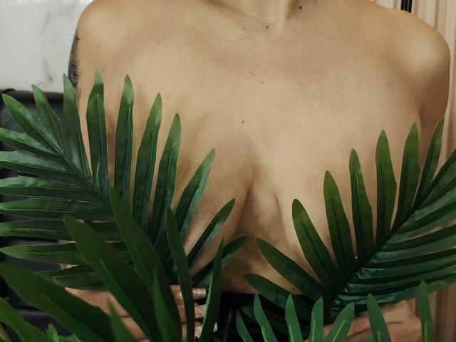 NenaBlack's tits are covered by green palm leaves, her tan skin and small tattoo on her right shoulder are visible. Her chest is prominently featured in the photo.