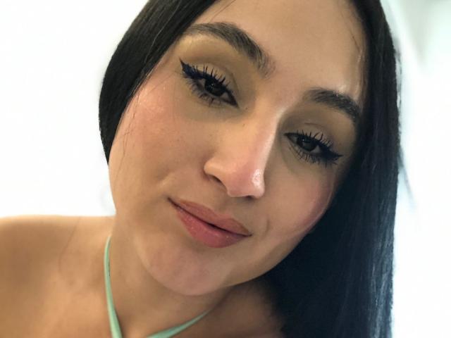 HiilaryDolce with long black hair, wearing a light green halter top, has thick black eyelashes, subtle pink lipstick, and a slight smile, looking directly at the camera.