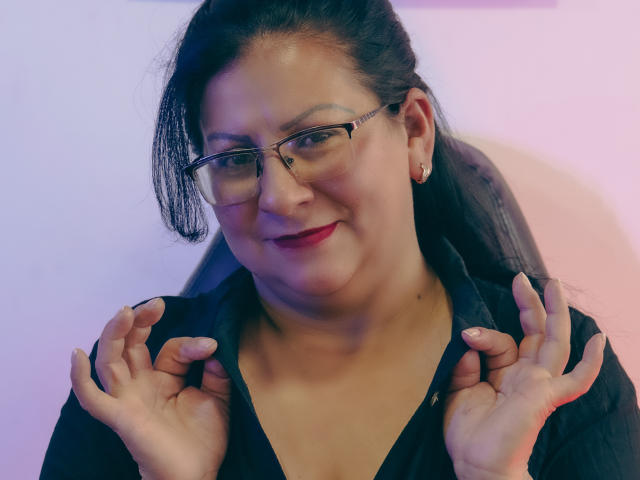 MissLadyy with glasses, dark hair in a ponytail, smiling, holding her shoulders with hands, wearing a black top, pink earbuds, and red lipstick.